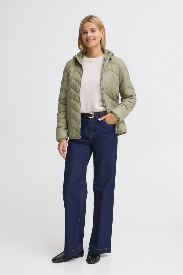 A woman modeling a light olive green quilted puffer jacket over a white top and navy blue wide-leg jeans, standing against a plain light gray studio background.