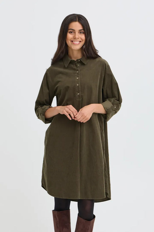 A woman modeling an olive green button-down shirt dress with long sleeves, paired with black tights and brown boots, against a light gray studio background.