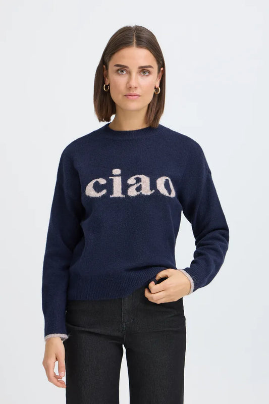 A woman wearing a navy blue knit sweater with the word 'ciao' knitted across the chest in a light beige tone, paired with black jeans, against a light gray studio background.