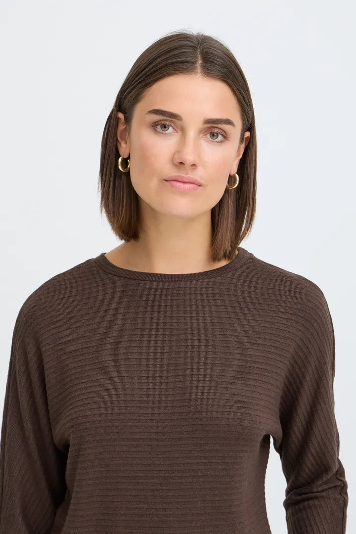 A woman with a straight brown bob haircut wearing a dark brown ribbed long-sleeve top and small gold hoop earrings, photographed against a light gray studio background.
