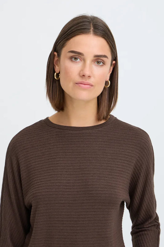 A woman with a straight brown bob haircut wearing a dark brown ribbed long-sleeve top and small gold hoop earrings, photographed against a light gray studio background.