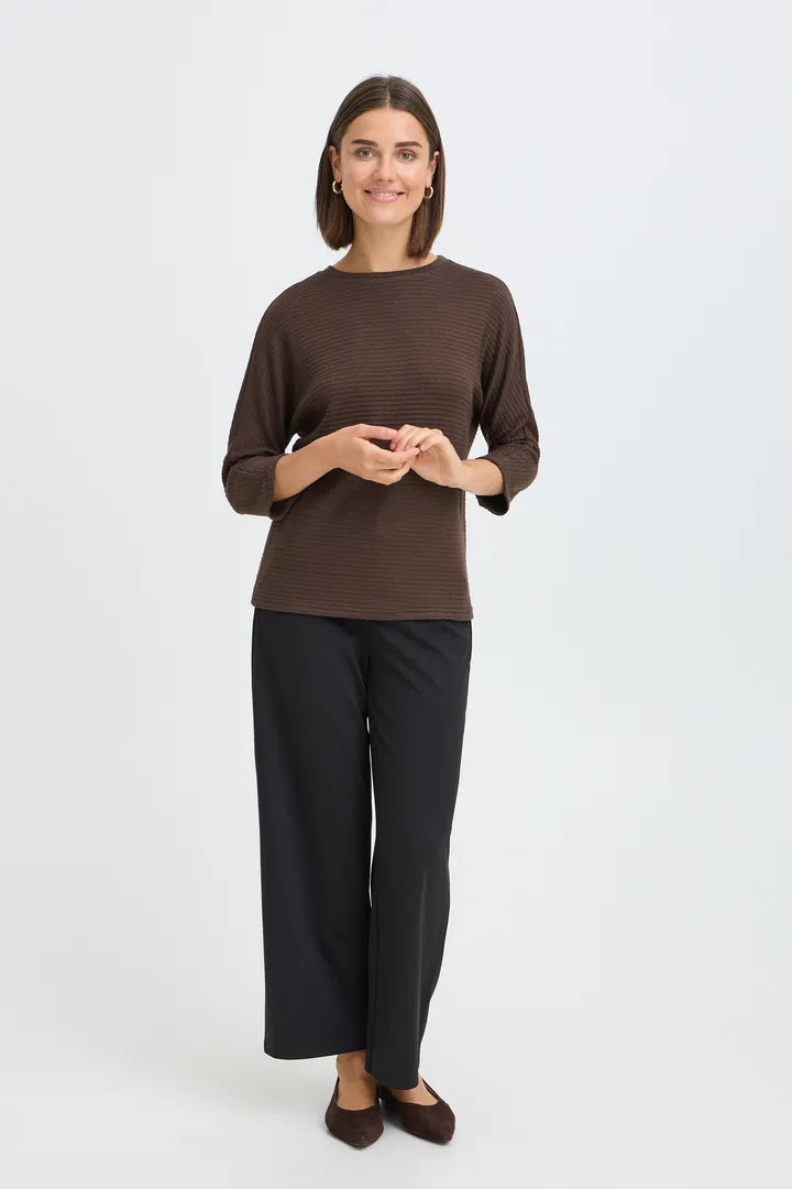A woman modeling a dark brown long-sleeve ribbed knit top with black wide-leg trousers and dark brown flats, standing against a light gray studio background.