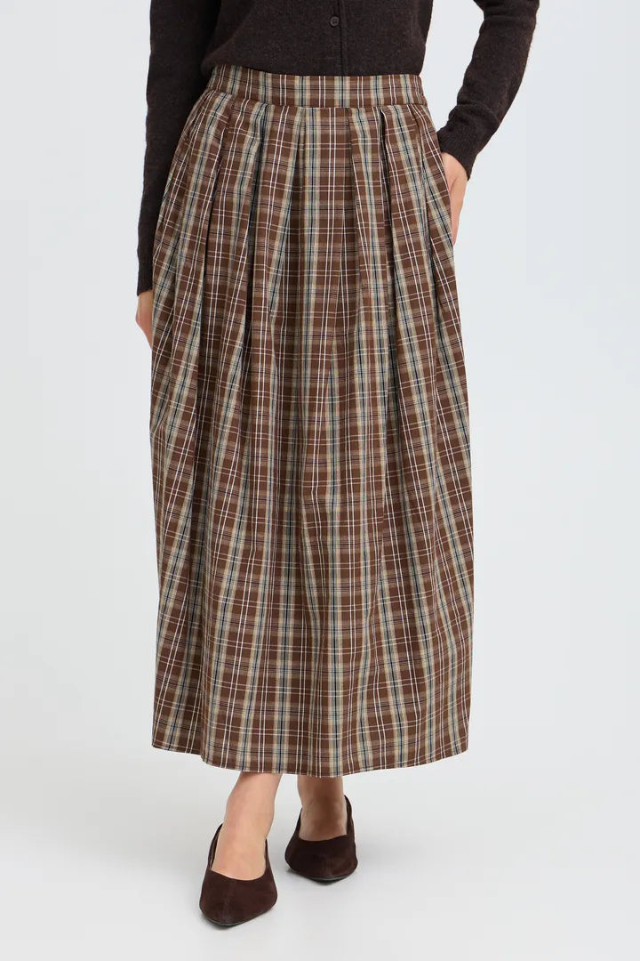 A model wearing a brown and beige plaid pleated midi skirt with a dark brown long-sleeve top and dark brown suede shoes, shown from the waist down against a light gray studio background.
