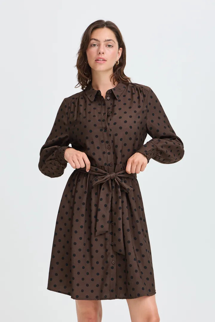 A woman modeling a dark brown long-sleeve shirt dress with a black polka dot pattern, featuring a button-up front, collar, and matching tie belt at the waist, set against a plain light gray background.