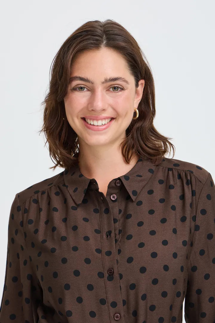 A woman with shoulder-length brown hair smiling, wearing a dark brown long-sleeve button-up blouse with black polka dots, set against a plain light gray background.
