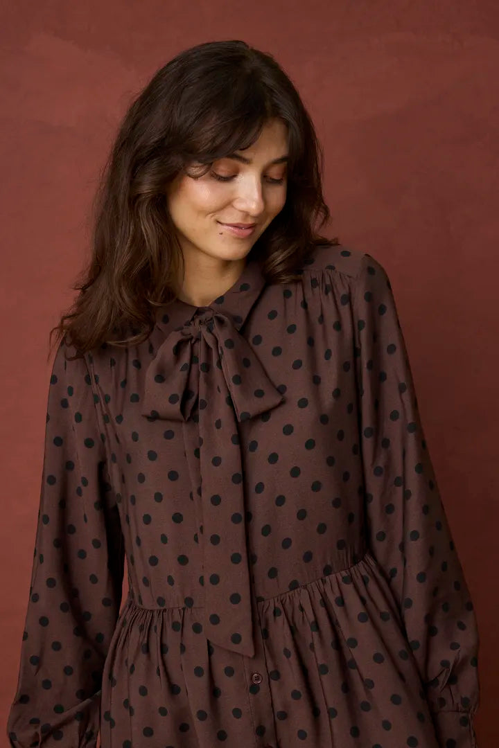 A woman wearing a brown long-sleeve blouse with black polka dots and a bow tie neckline, standing against a solid dark reddish-brown background.