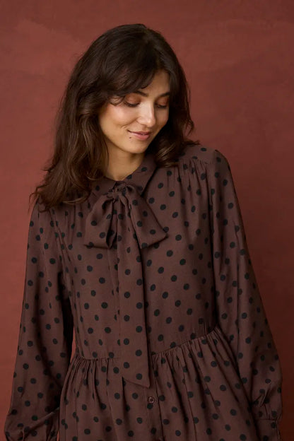 A woman wearing a brown long-sleeve blouse with black polka dots and a bow tie neckline, standing against a solid dark reddish-brown background.