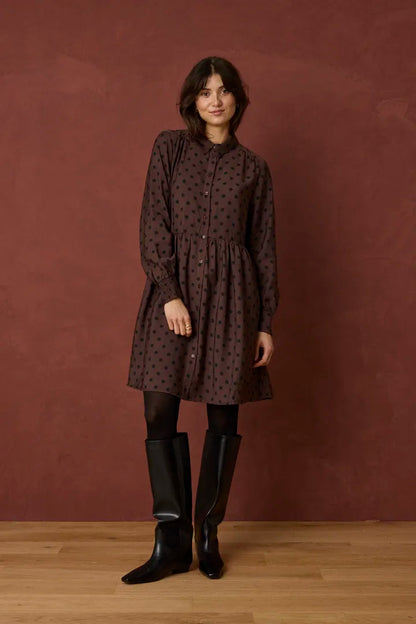 A woman stands against a textured reddish-brown wall on a light wood floor, wearing a dark brown long-sleeve button-front dress with a small repeating pattern, black tights, and tall black boots.
