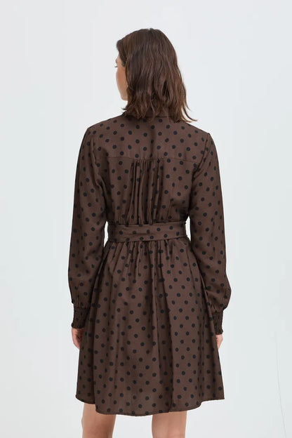 A woman viewed from the back wearing a brown long-sleeve dress with a black polka dot pattern and a matching tie belt at the waist, set against a plain white studio background.