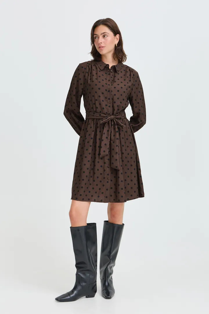 A woman modeling a dark brown long-sleeve mini dress with a black polka dot pattern, a pointed collar, and a matching waist tie, paired with black knee-high boots against a light gray studio background.