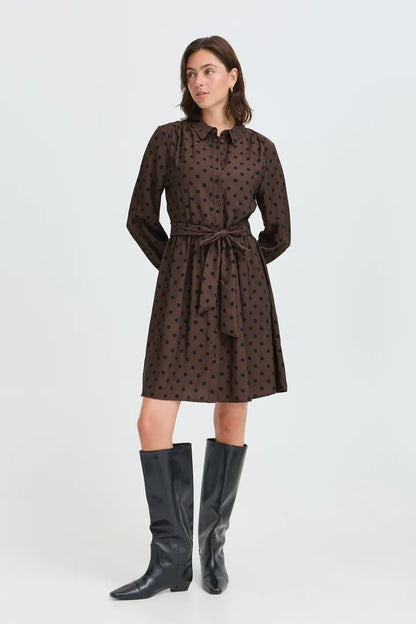 A woman modeling a dark brown long-sleeve mini dress with a black polka dot pattern, a pointed collar, and a matching waist tie, paired with black knee-high boots against a light gray studio background.
