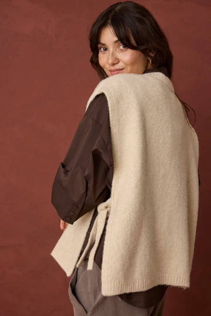 A woman wearing a cream-colored knit sleeveless vest over a dark brown jacket, photographed against a textured reddish-brown studio backdrop.