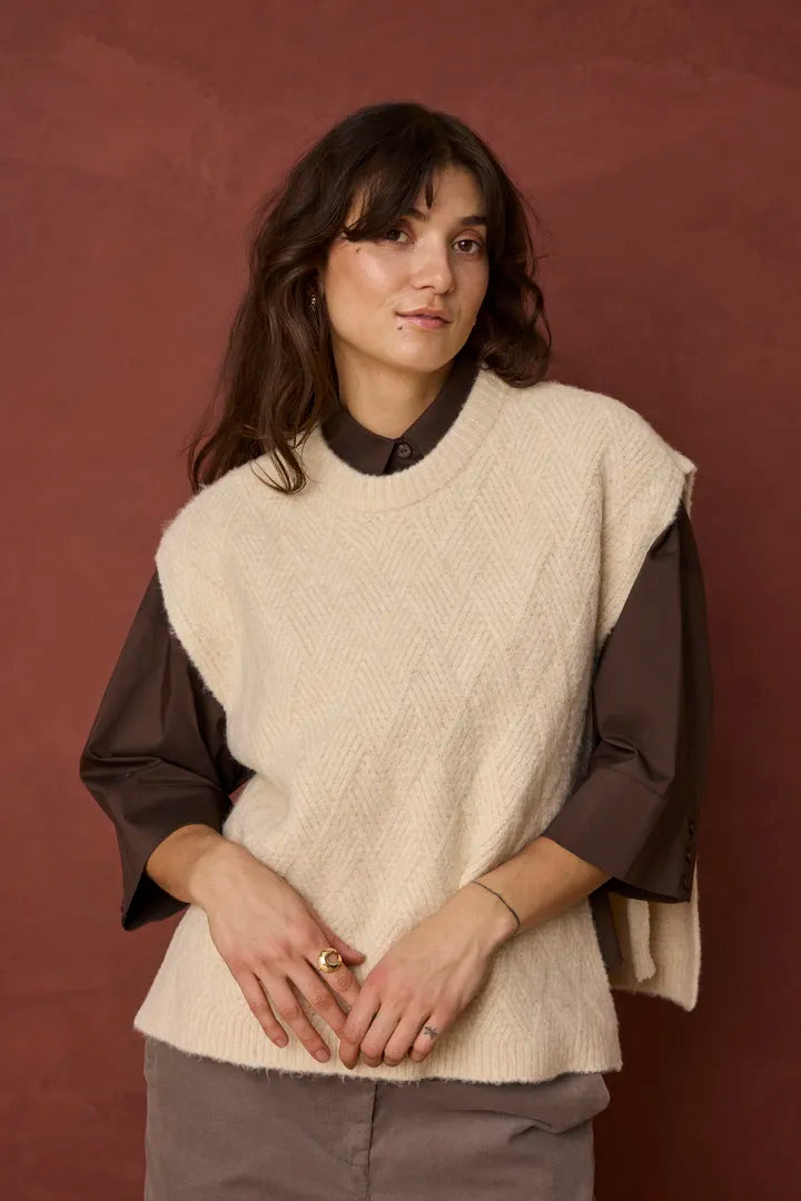 A woman wearing a cream-colored textured knit sweater vest over a dark brown long-sleeve shirt, standing against a textured reddish-brown background.