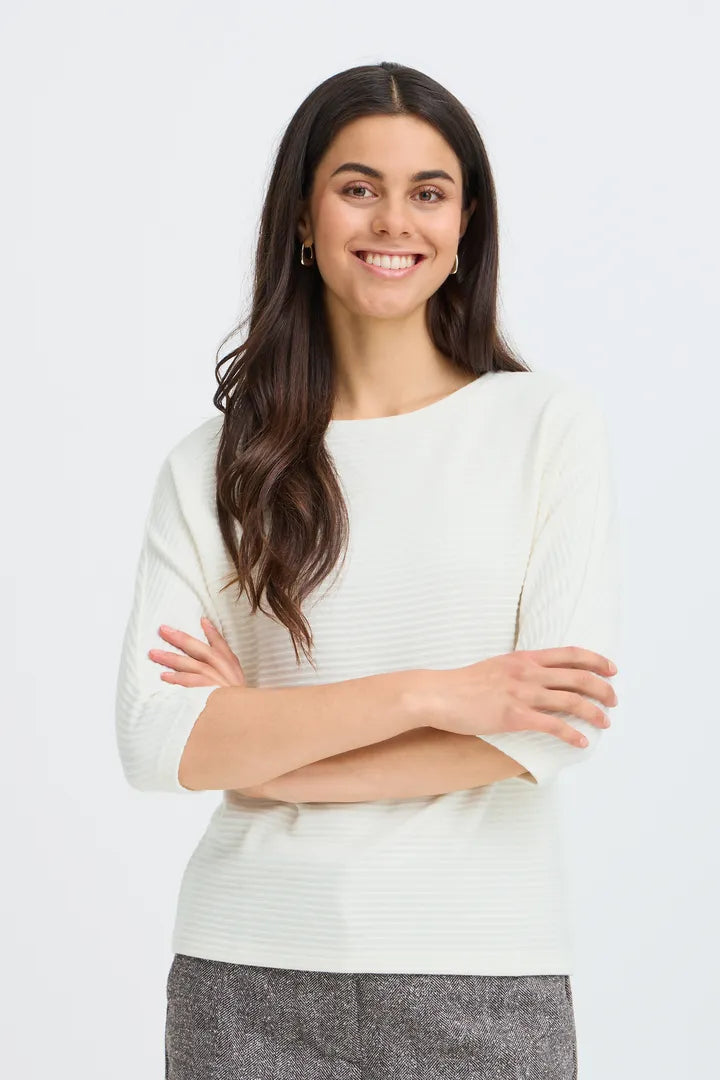 A smiling woman with long dark hair wearing a cream ribbed knit sweater and gray pants, standing with arms crossed against a light gray studio background.