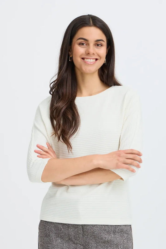 A smiling woman with long dark hair wearing a cream ribbed knit sweater and gray pants, standing with arms crossed against a light gray studio background.