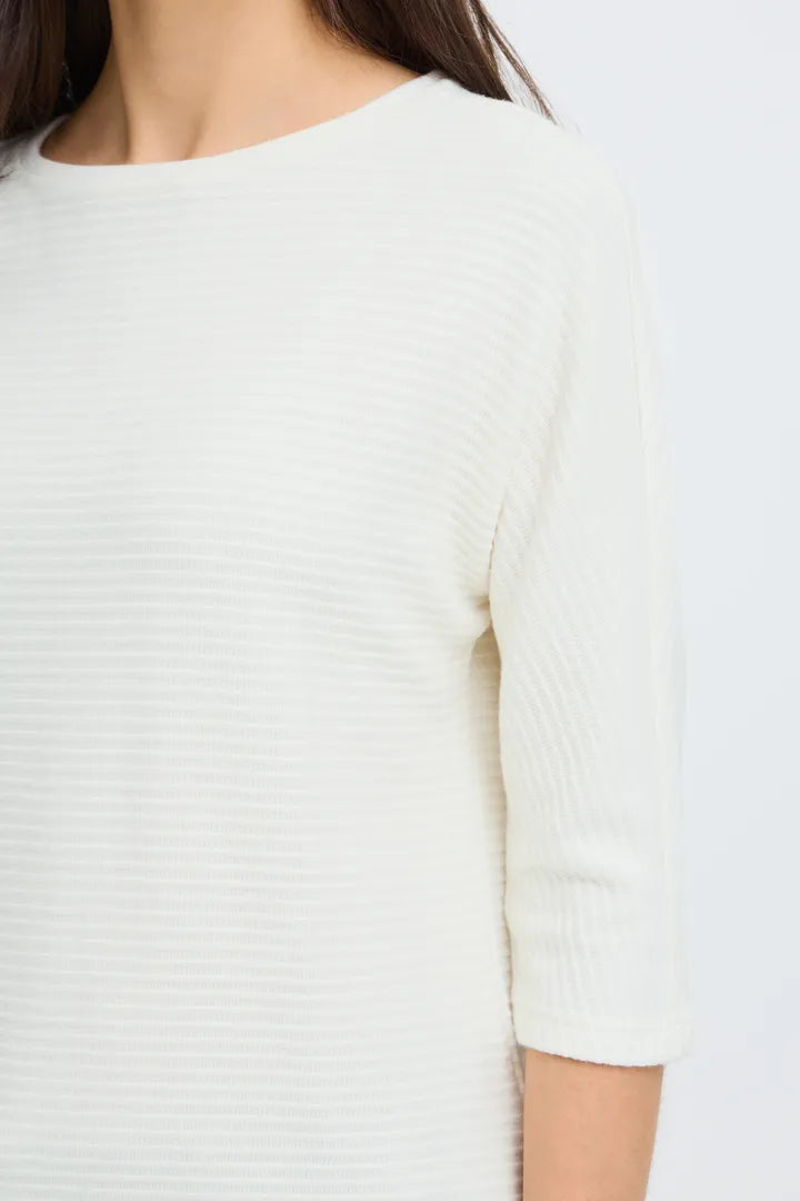 Close-up of a person wearing a ribbed knit top in off-white or cream, showing the chest, shoulder, and elbow area against a plain light background.