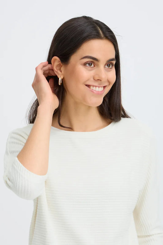 A smiling woman with dark hair wearing a white ribbed long-sleeve top and a small gold hoop earring, shown against a light gray studio background.