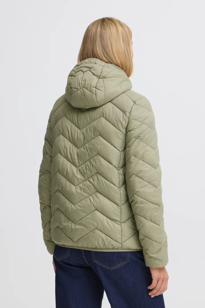 A woman viewed from the back wearing a light olive green quilted puffer jacket with a hood and chevron stitching, paired with dark blue jeans, against a plain light gray background.