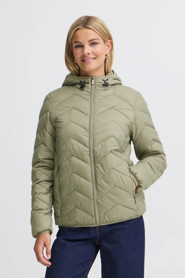 A woman wearing a light olive green hooded puffer jacket with a chevron quilted pattern and navy blue jeans, photographed against a plain light gray studio background.