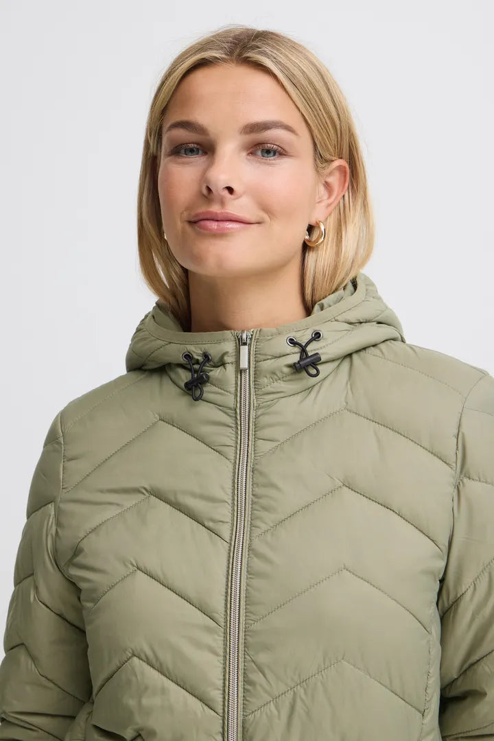 A woman wearing a light olive green hooded puffer jacket with a front zipper and black drawstrings, shown against a plain light gray background.