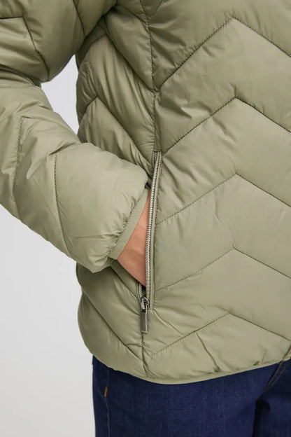 Close-up of a person wearing a quilted puffer jacket in a muted sage green color, with a hand sliding into the side pocket. The jacket features a chevron quilting pattern and a silver zipper.