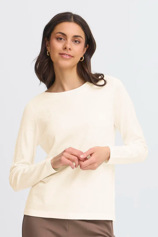 A woman wearing a cream-colored long-sleeve knit top with a boat neckline, paired with brown pants, against a light gray studio background.