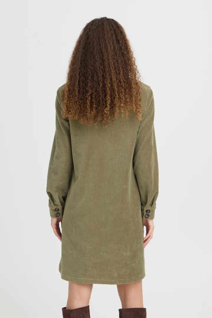 A model viewed from the back wearing a sage green corduroy tunic dress with long sleeves and button cuffs, paired with brown boots, against a plain light gray background.