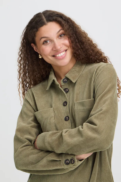 A smiling woman with curly brown hair wearing an olive green corduroy button-up shirt, posing with her arms crossed against a light gray background.