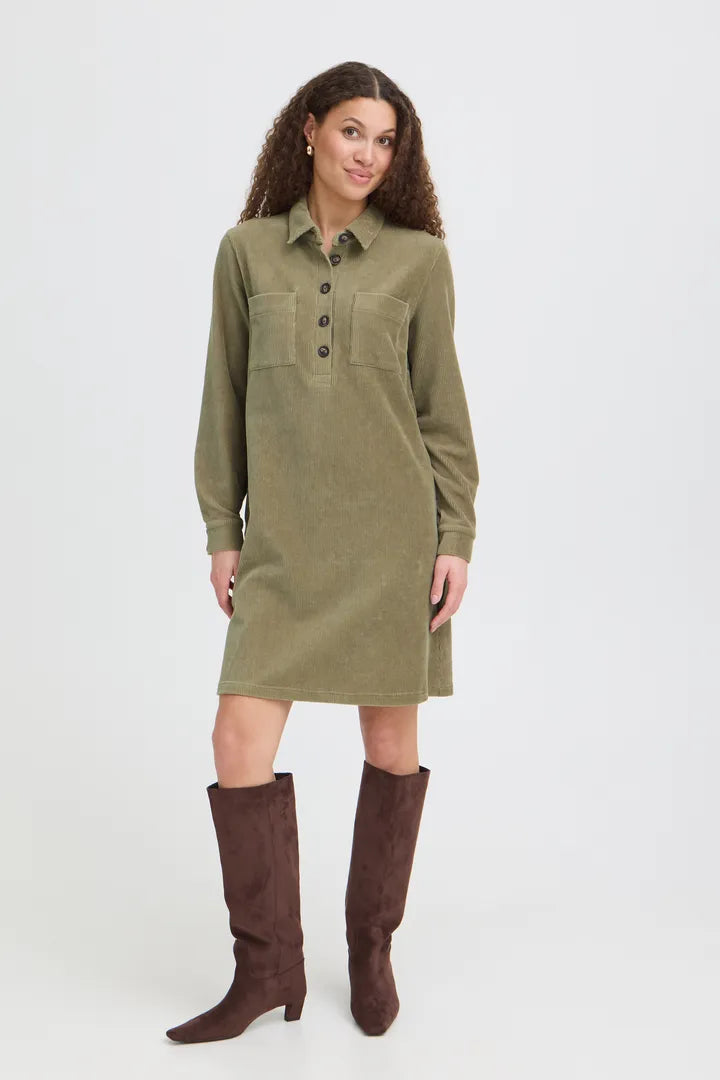 A woman modeling a long-sleeve olive green corduroy shirt dress with a buttoned collar and chest pockets, paired with dark brown knee-high boots, against a light gray studio background.