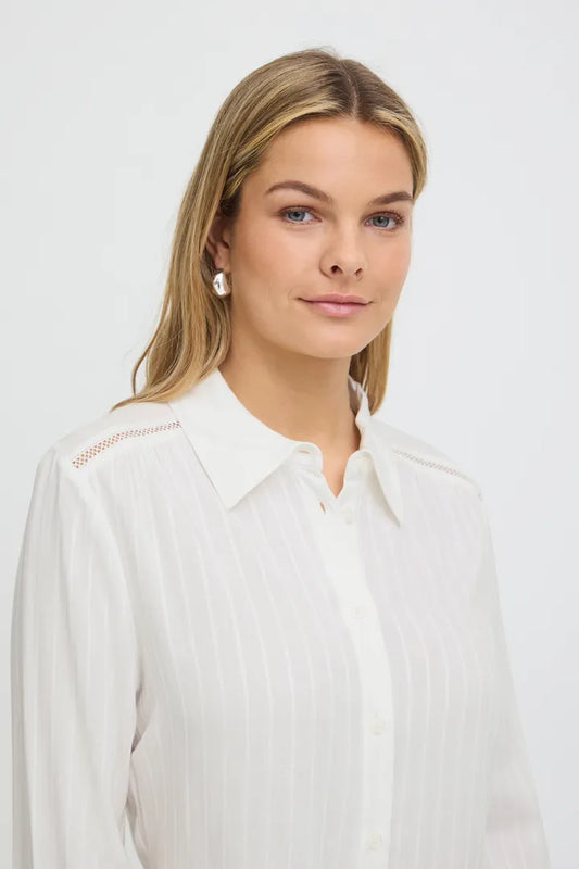 A woman with blonde hair wearing a white long-sleeve button-up blouse with subtle vertical pleats and decorative stitching on the shoulder, photographed against a light gray studio background.