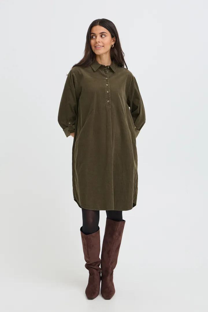 A woman modeling an olive green corduroy shirt dress with a button-down front, paired with dark tights and brown knee-high boots, against a plain light gray studio background.