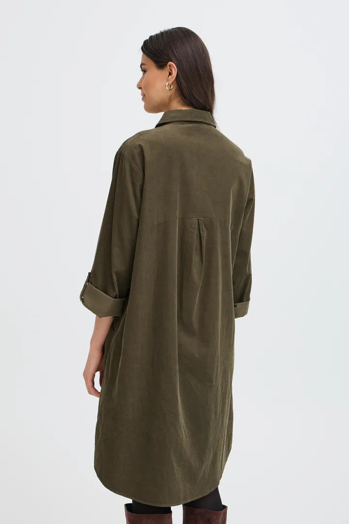 A woman shown from the back wearing an olive green corduroy shirt coat with rolled-up sleeves, photographed against a plain light gray studio background.