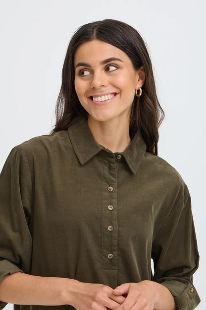 A woman with dark hair wearing an olive green button-down shirt, shown from the chest up against a light gray studio background.