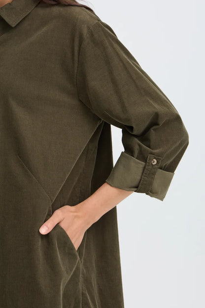 Close-up of a person wearing an olive green long-sleeve shirt dress with rolled-up cuffs and a side pocket, shown against a light gray background.