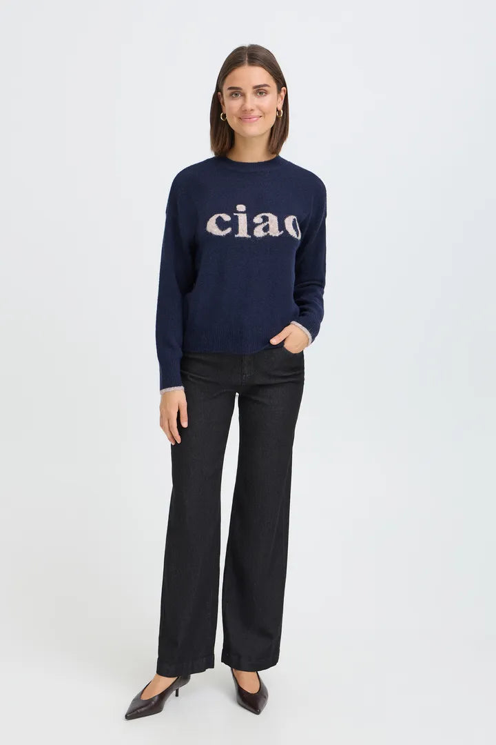 A woman modeling a navy blue crewneck sweater with 'ciao' written across the chest in a light beige serif font, paired with black wide-leg trousers and black pointed-toe heels, against a plain light gray studio background.