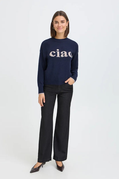 A woman modeling a navy blue crewneck sweater with 'ciao' written across the chest in a light beige serif font, paired with black wide-leg trousers and black pointed-toe heels, against a plain light gray studio background.
