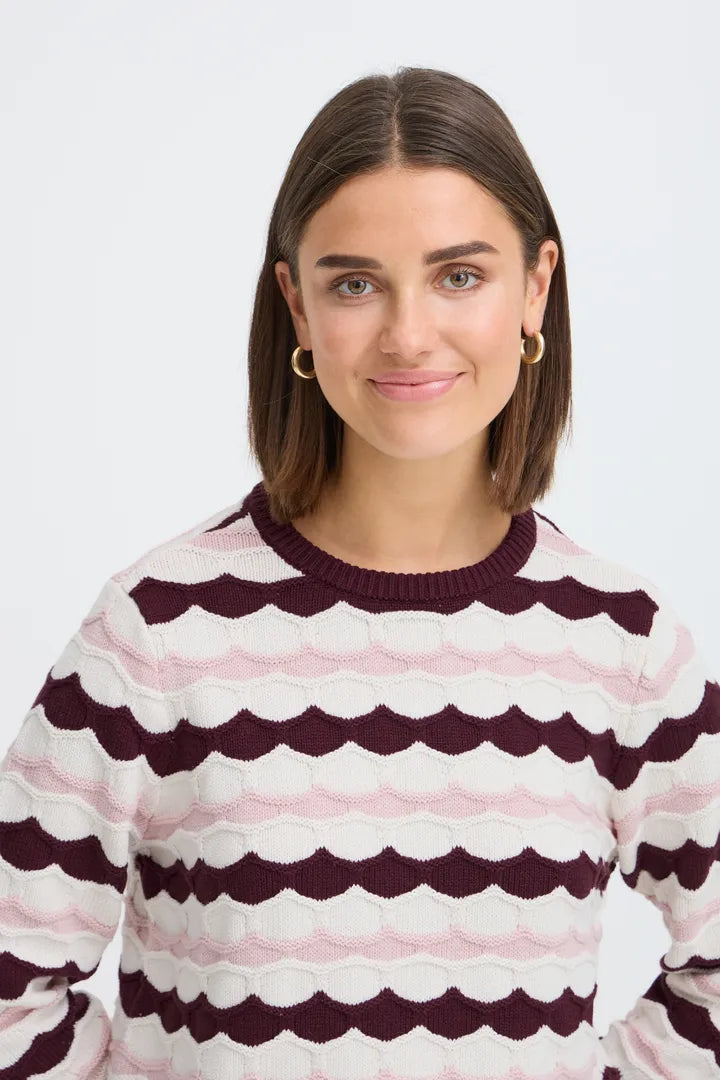 A woman with shoulder-length brown hair wearing a knit crewneck sweater with horizontal wavy stripes in dark burgundy and light pink on an off-white base, photographed against a clean light gray background.