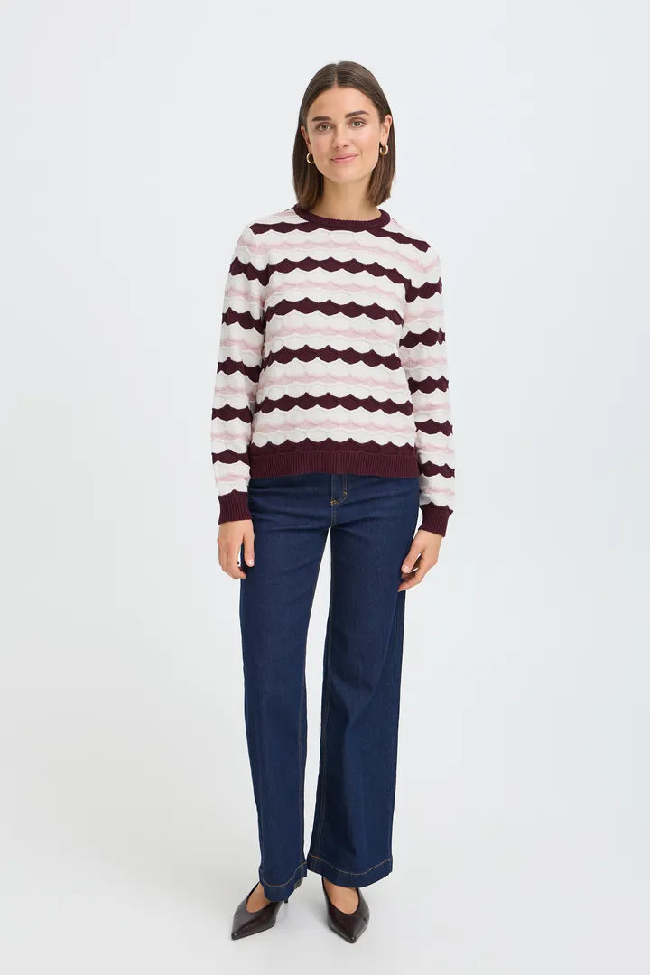A woman modeling a long-sleeve knit sweater with horizontal wavy stripes in cream and burgundy, paired with dark blue jeans and dark brown pointed flats, against a plain white studio background.