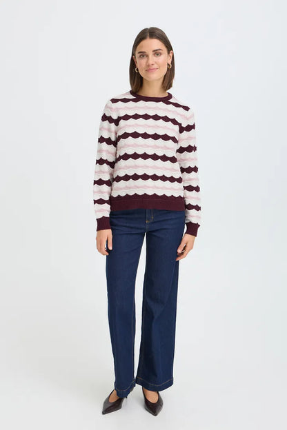A woman modeling a long-sleeve knit sweater with horizontal wavy stripes in cream and burgundy, paired with dark blue jeans and dark brown pointed flats, against a plain white studio background.