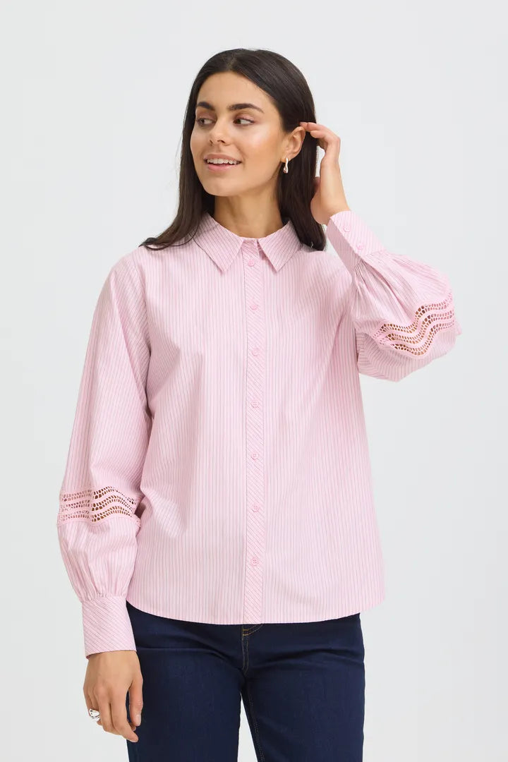A woman modeling a light pink and white vertically striped long-sleeve blouse with lace trim on the cuffs, paired with dark blue jeans, against a light gray studio background.