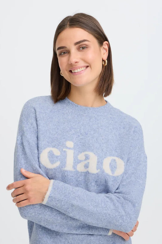 A smiling woman with shoulder-length brown hair wearing a light blue knit sweater with the word 'ciao' in large cream letters across the chest, posed against a plain light gray background.