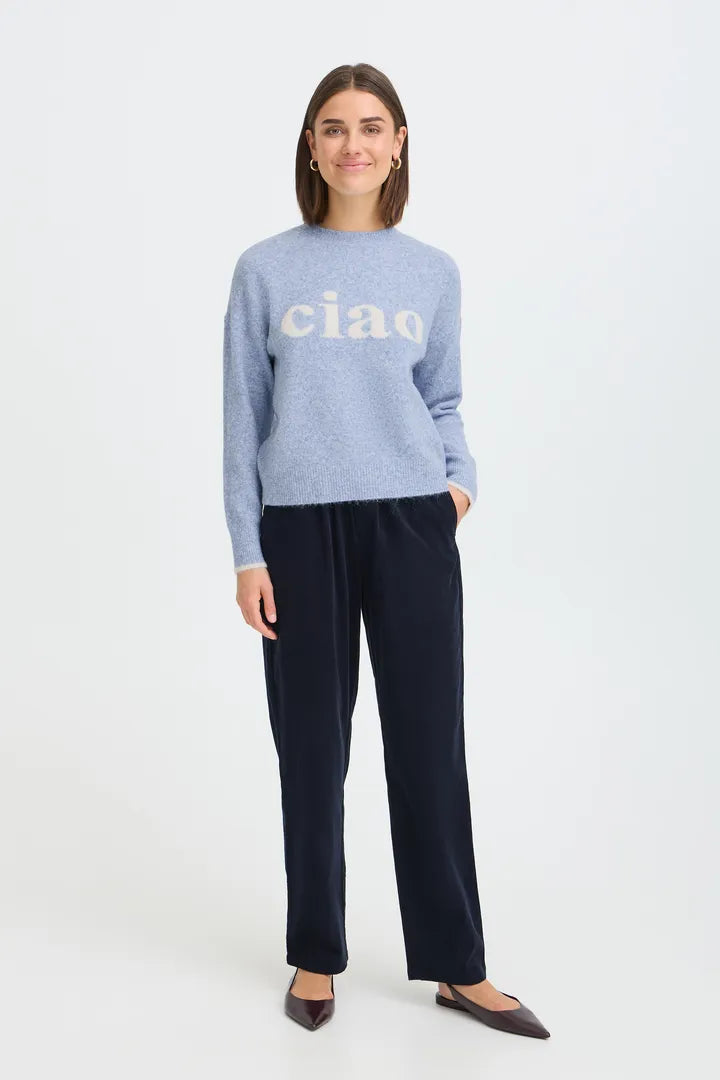 A woman modeling a light blue knit sweater with 'ciao' printed in white across the chest, paired with dark navy wide-leg trousers and dark brown pointed flats, against a plain white studio background.