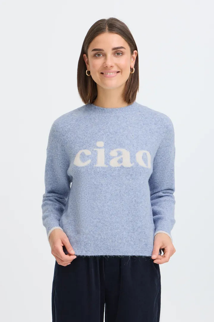 A woman wearing a light blue knit sweater with the word 'ciao' in white across the chest, paired with dark navy pants, photographed against a plain light gray studio background.