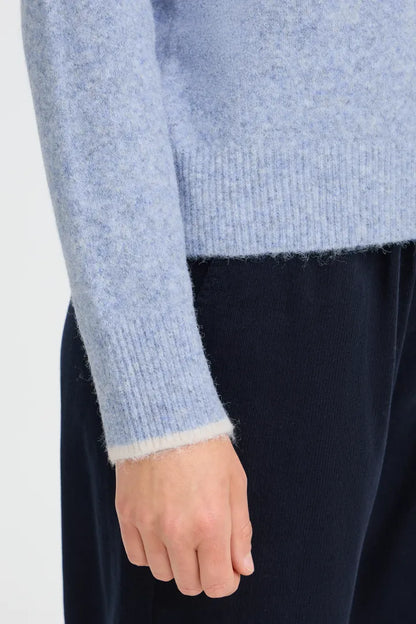 Close-up of a person wearing a light blue fuzzy knit sweater with ribbed cuffs and hem, paired with dark navy pants, against a plain light background.