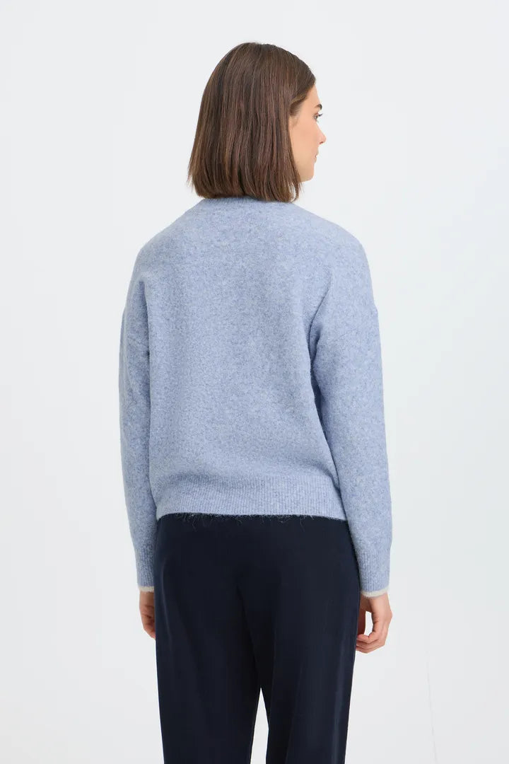 A woman viewed from the back wearing a light blue knit sweater and dark navy trousers, standing against a plain white studio background.