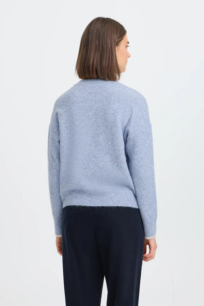 A woman viewed from the back wearing a light blue knit sweater and dark navy trousers, standing against a plain white studio background.
