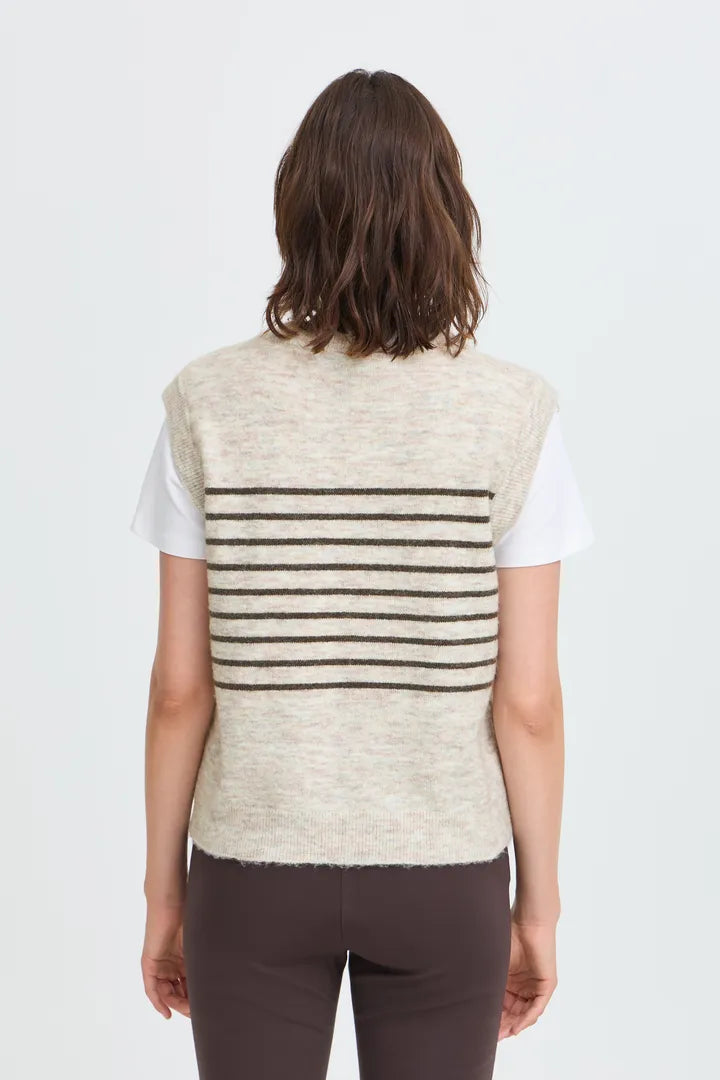A woman viewed from behind wearing a cream-colored knit sweater vest with horizontal dark brown stripes over a white t-shirt and dark brown pants, against a light gray studio background.