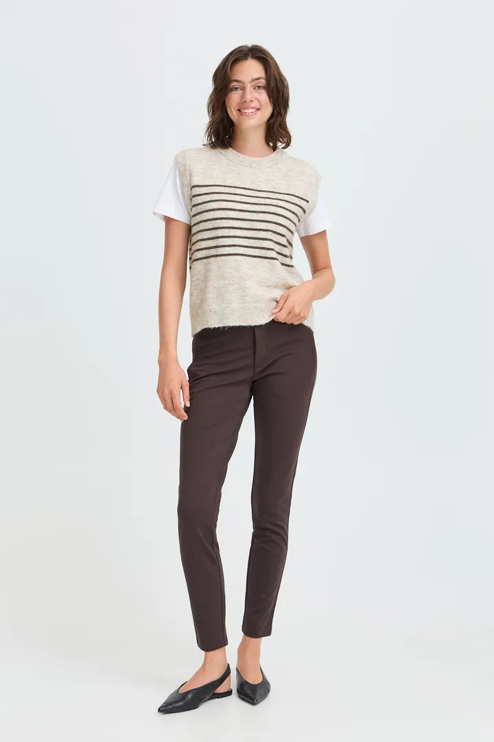 A woman modeling a short-sleeve knit top with horizontal stripes over a white tee, paired with dark brown pants and black flats, against a light gray studio background.