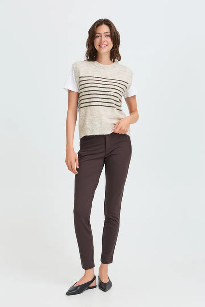 A woman modeling a short-sleeve knit top with horizontal stripes over a white tee, paired with dark brown pants and black flats, against a light gray studio background.