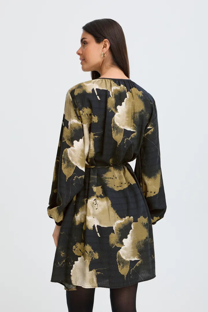 A woman shown from the back wearing a long-sleeve tunic dress with a black base and a large abstract floral print in beige and olive green, paired with black tights against a light gray studio background.